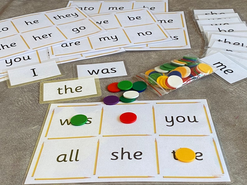 May include: A game board with nine squares, each containing a word. The words are "was", "the", "you", "all", "she", "toe", and "me". There are also colorful chips on the board and in a plastic bag.