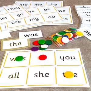May include: A game board with a grid of nine squares, each containing a word. The words are "was", "the", "you", "all", "she", "toe", "ws", and two blank squares. There are colorful counters on the board and in a plastic bag.