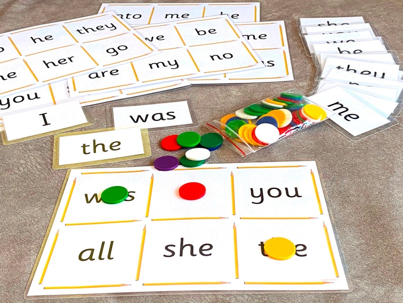 May include: A game board with a grid of nine squares, each containing a word. The words are "was", "the", "you", "all", "she", "toe", "ws", and two blank squares. There are colorful counters on the board and in a plastic bag.