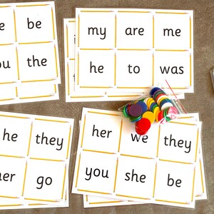 May include: A set of white cards with sight words printed on them, such as "I", "me", "be", "was", "you", "the", "he", "to", "into", "she", "her", "go", "we", "they", "my", "are", "her", "you", "be", "no", "lo". The cards are arranged in a grid pattern and are surrounded by a yellow border. There are also a few colorful counters scattered around the cards.