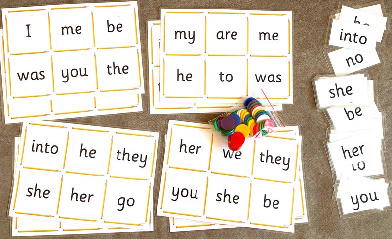 May include: A set of white cards with sight words printed on them, such as "I", "me", "be", "was", "you", "the", "he", "to", "into", "she", "her", "go", "we", "they", "my", "are", "her", "you", "be", "no", "lo". The cards are arranged in a grid pattern and are surrounded by a yellow border. There are also a few colorful counters scattered around the cards.