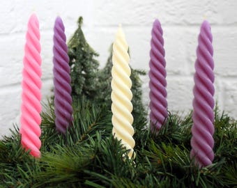 May include: Five twisted candles in a wreath of evergreen branches. The candles are pink, purple, and cream.