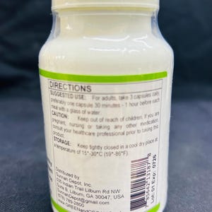 Op de afbeelding: Een witte plastic fles met een witte dop, met capsules. Het etiket bevat gebruiksinstructies, waarschuwingen en bewaarinstructies. De fles is voorzien van de informatie en website van de distributeur.