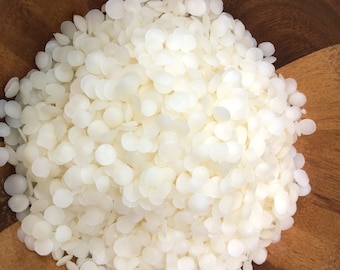 May include: A close-up view of a bowl filled with white wax flakes. The flakes are small and round, and they are piled high in the bowl.