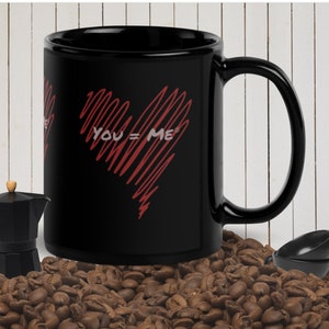 Può includere: Tazza da caffè nera con un design a forma di cuore rosso e il testo "YOU = ME" su di essa. La tazza è appoggiata su un letto di chicchi di caffè.