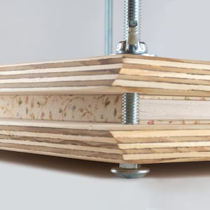 May include: A wooden book press with a layered plywood construction. The press features a central space for holding items, secured by a threaded rod and metal hardware. The wood has a natural, light brown color.
