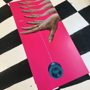 May include: A hand with a ring on the finger holds a string attached to a dark blue yo-yo on a bright pink background.