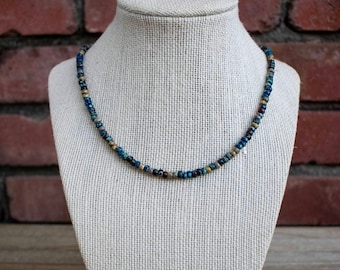 May include: A blue and gold beaded necklace on a fabric-covered mannequin. The necklace is made of small, round beads in shades of blue, gold, and brown.