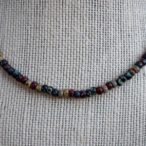 May include: A necklace made of small, colorful beads. The beads are brown, red, and blue with white speckles. The necklace is on a beige fabric background.