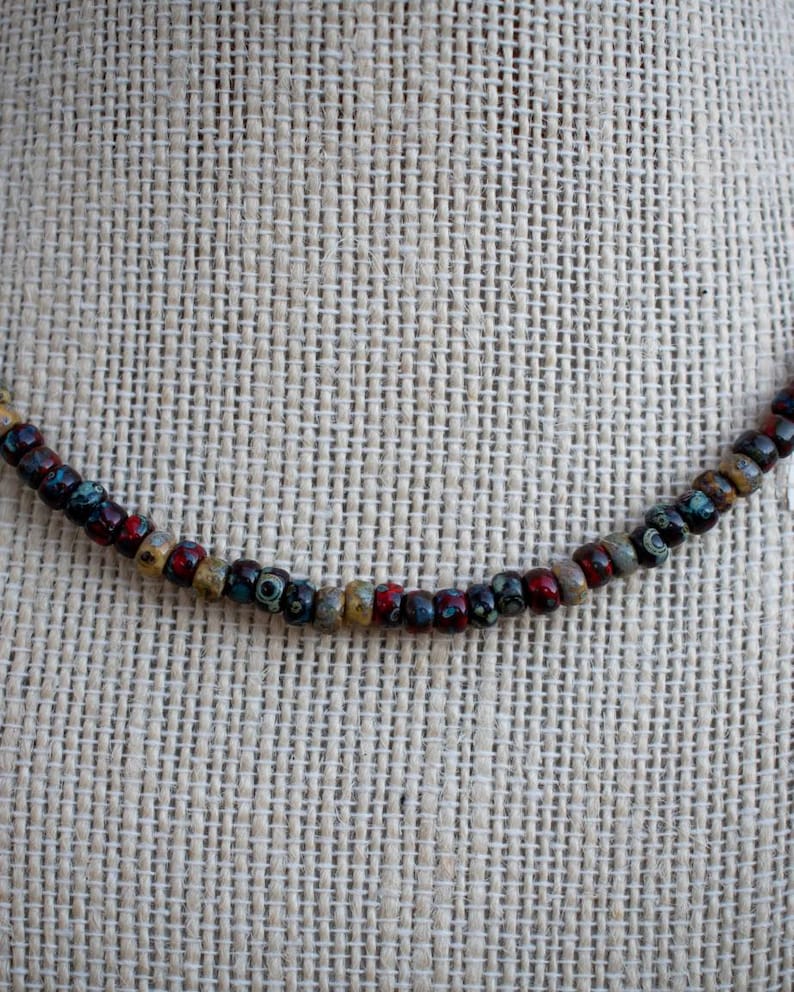 May include: A necklace made of small, colorful beads. The beads are brown, red, and blue with white speckles. The necklace is on a beige fabric background.