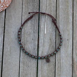 May include: A brown macrame necklace with turquoise-colored beads. The necklace is on a wooden surface. The beads are rectangular and evenly spaced along the cord. The necklace is adjustable with a sliding knot closure.