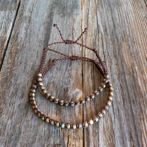 May include: Two beaded bracelets with brown cord, resting on a weathered wooden surface. The beads are a mix of gold, green, and cream colors. The bracelets are adjustable with a sliding knot closure.