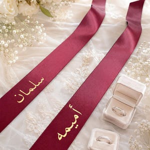 May include: Two long burgundy satin ribbons featuring gold Arabic calligraphy text, accompanied by two small velvet ring boxes, each containing a silver-toned ring. The ribbons display the names Salman and Amima in a metallic gold finish.