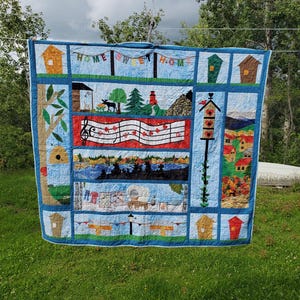 Peut inclure: Une courtepointe en patchwork à plusieurs panneaux présentant diverses scènes appliquées, notamment des nichoirs, une portée musicale avec des feuilles d'érable rouges, un paysage avec un élan, un chariot couvert et le texte HOME SWEET HOME.