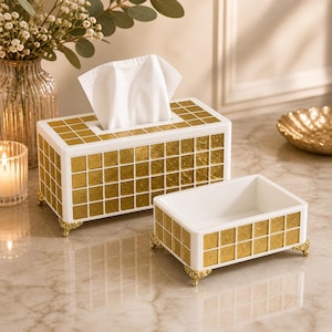 Op de afbeelding: Een witte en gouden mozaïek tissuebox en een bijpassende kleine container. De tissuebox heeft een rechthoekige vorm met een witte bovenkant en gouden mozaïektegels aan de zijkanten. Beide items hebben sierlijke gouden voetjes.
