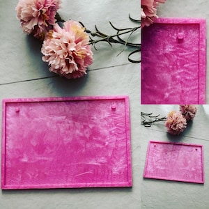 Pode incluir: Duas bandejas de exibição de joias de veludo rosa com uma borda elevada e dois pequenos orifícios no topo. As bandejas estão sobre uma superfície branca.