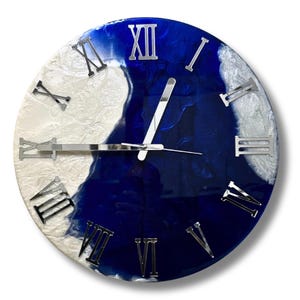 Könnte beinhalten: Runde Wanduhr mit zweifarbigem Design. Das Zifferblatt ist in weiße und tiefblaue Abschnitte mit Marmoreffekt unterteilt. Silberne römische Ziffern markieren die Stunden. Die Uhrzeiger sind silberfarben.