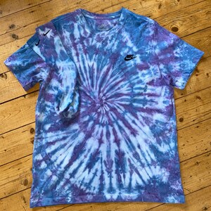 Op de afbeelding: Een blauw en paars tie-dye T-shirt met een zwart Nike-logo op de linkerborst. Het shirt is gevouwen en ligt plat op een houten oppervlak.