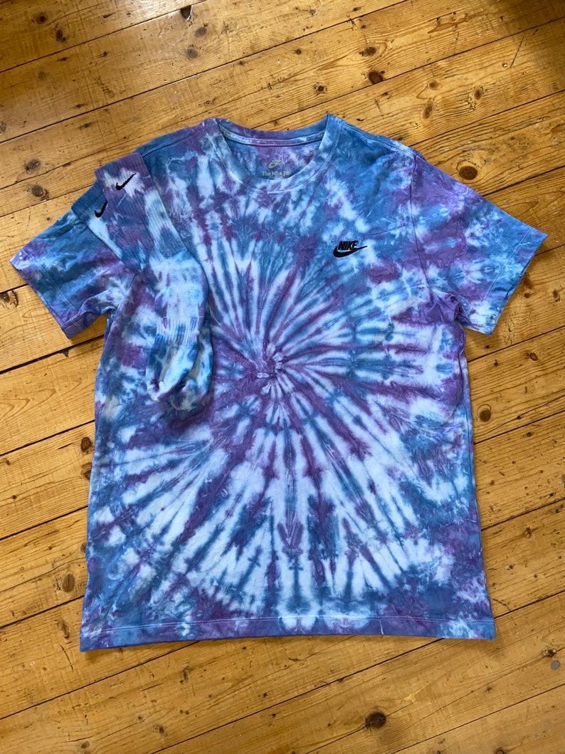 Op de afbeelding: Een blauw en paars tie-dye T-shirt met een zwart Nike-logo op de linkerborst. Het shirt is gevouwen en ligt plat op een houten oppervlak.