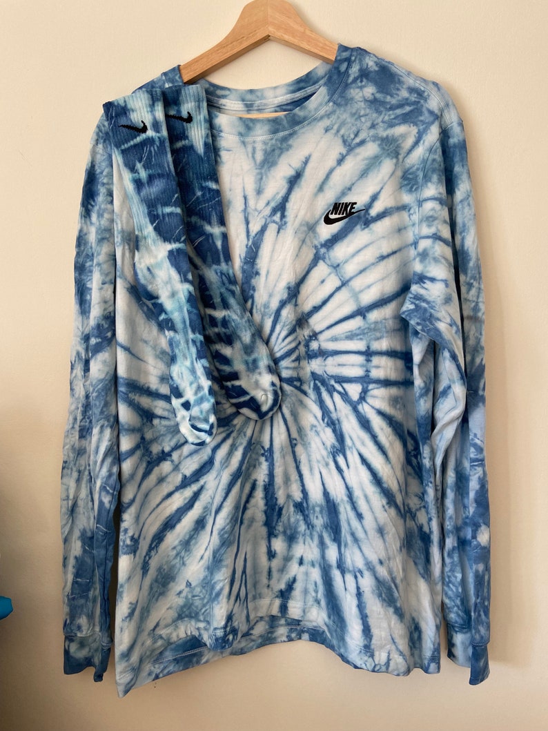 Op de afbeelding: Een blauw tie-dye longsleeve shirt met een wit Nike logo op de linker borst. Het shirt wordt gecombineerd met een paar blauwe tie-dye sokken.