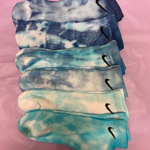 May include: Six pairs of tie-dye socks with a black Nike swoosh logo. The socks are in shades of blue and white.