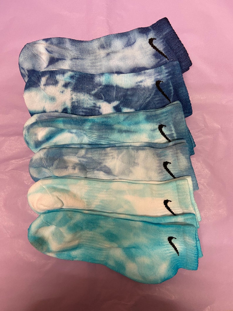 May include: Six pairs of tie-dye socks with a black Nike swoosh logo. The socks are in shades of blue and white.