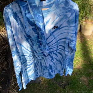 Op de afbeelding: Een blauw tie-dye longsleeve shirt met een wit Nike logo op de voorkant. Het shirt hangt aan een hanger en heeft een blauw en wit tie-dye patroon.