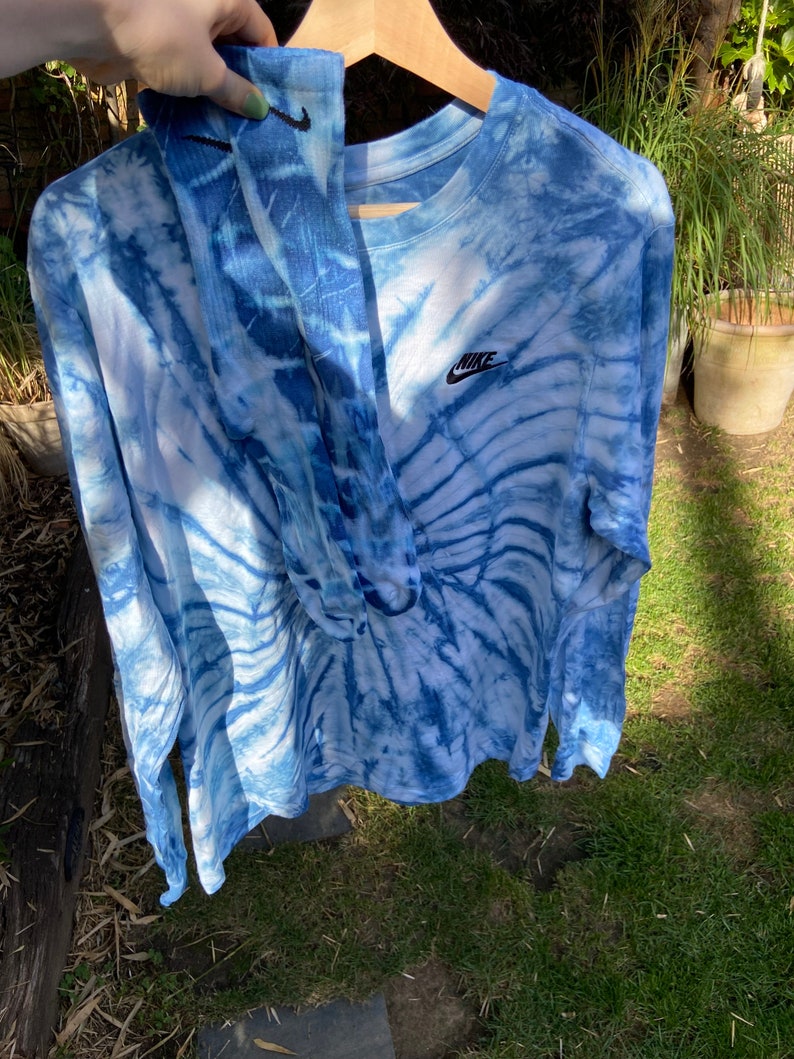Op de afbeelding: Een blauw tie-dye longsleeve shirt met een wit Nike logo op de voorkant. Het shirt hangt aan een hanger en heeft een blauw en wit tie-dye patroon.