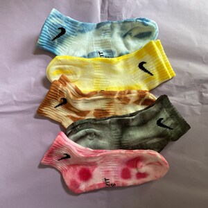 May include: Five pairs of colorful tie-dye socks with a black Nike logo. The socks are stacked on top of each other, with the blue sock on top and the pink sock on the bottom. The socks are all different colors, including blue, yellow, brown, gray, and pink.