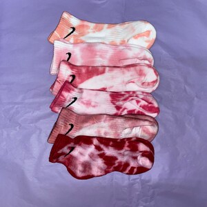 May include: A stack of six pairs of pink and white tie-dye socks with a black Nike swoosh logo on each sock.