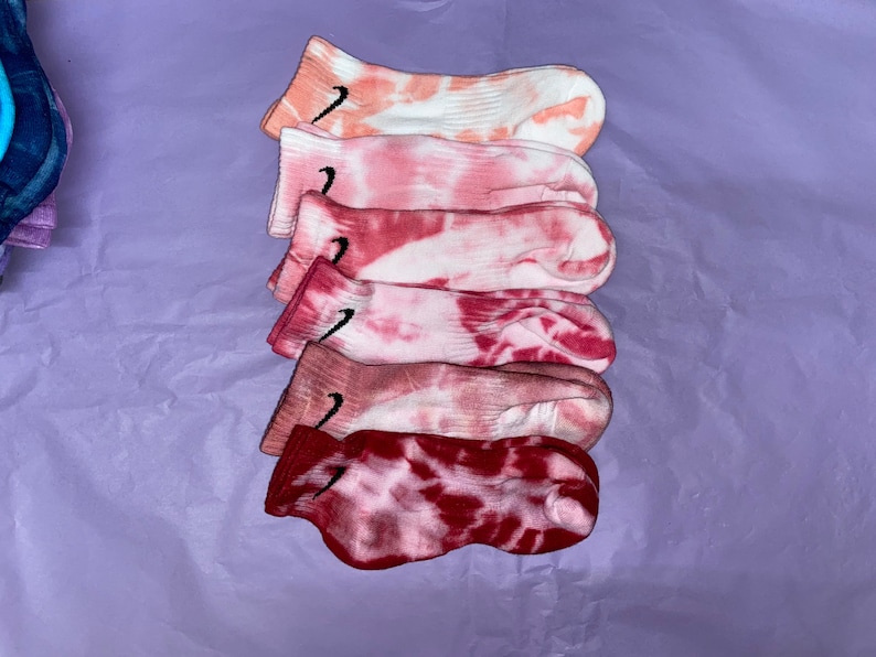 May include: A stack of six pairs of pink and white tie-dye socks with a black Nike swoosh logo on each sock.