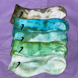 May include: Five pairs of tie-dye socks in various shades of green and blue. The socks have a white background with a black Nike swoosh logo on each sock.