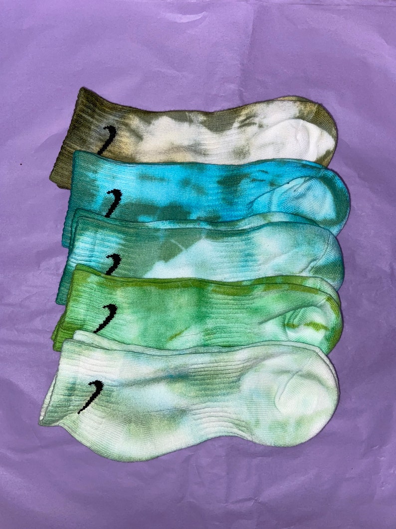 May include: Five pairs of tie-dye socks in various shades of green and blue. The socks have a white background with a black Nike swoosh logo on each sock.