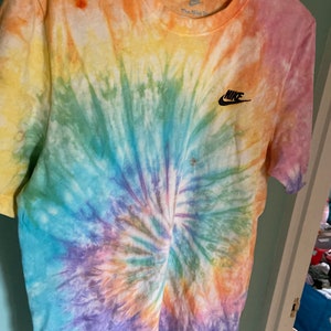 Op de afbeelding: Een tie-dye T-shirt met een regenboogspiraalpatroon. Het shirt heeft een zwart Nike-logo op de linkerborst en de tekst "The Nike" op het label.