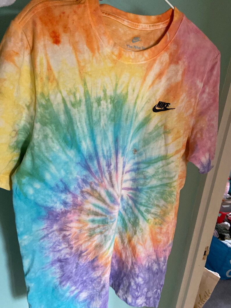 Op de afbeelding: Een tie-dye T-shirt met een regenboogspiraalpatroon. Het shirt heeft een zwart Nike-logo op de linkerborst en de tekst "The Nike" op het label.