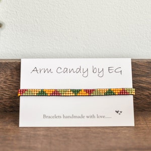 Könnte beinhalten: Ein farbenfrohes Armband mit grünen, gelben, roten und braunen Perlen. Das Armband wird auf einer weißen Karte mit dem Text "Arm Candy by EG" und "Bracelets handmade with love..." präsentiert.