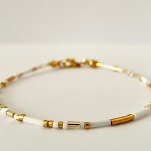 May include: A delicate white and gold beaded bracelet with a simple clasp closure. The bracelet features small, round white beads interspersed with gold beads, creating a minimalist and elegant look.