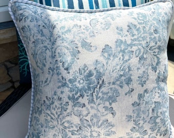 May include: Square throw pillow with a light blue floral pattern on a cream background. The pillow has a white rope trim and is placed in front of a striped pillow. The floral design is a classic damask style.
