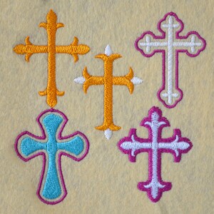 May include: Five embroidered crosses in different colors and styles. The crosses are orange, white, turquoise, and purple.