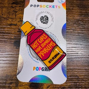 May include: A PopSockets PopGrip featuring a red and yellow bottle design with the text "HOT GIRLS HAVE STOMACH PROBLEMS" and "12 FL OZ (354ml)". The PopGrip is on a white and silver backing with the PopSockets and Mosstera Creations logos.
