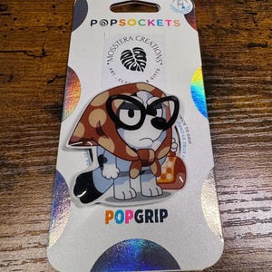 May include: A PopSockets PopGrip featuring a cartoon dog character wearing glasses and a headscarf, holding a purse. The PopGrip is attached to a white and iridescent backing card with the PopSockets and Mossstera Creations logos.
