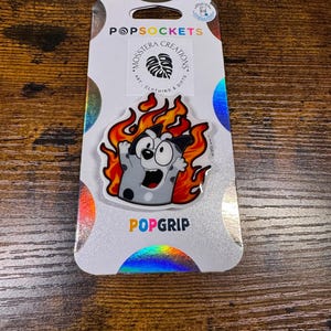 May include: A PopSocket featuring a cartoon character engulfed in flames. The character is gray with black spots and has a surprised expression. The PopSocket is on a white backing with the words "PopSockets" and "PopGrip".