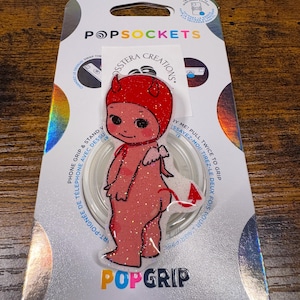 May include: A PopSockets PopGrip featuring a glittery red devil character with a red hat, wings, and tail. The PopGrip is attached to a silver and white backing card with the PopSockets and PopGrip logos, and the text "SSTERA CREATIONS."
