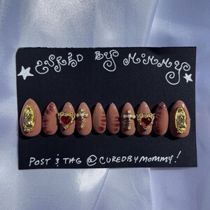 May include: A set of almond-shaped, press-on nails in a nude color, adorned with gold religious charms, red heart-shaped gems, and a marbled design. The black card reads "Cured by Mommy!"