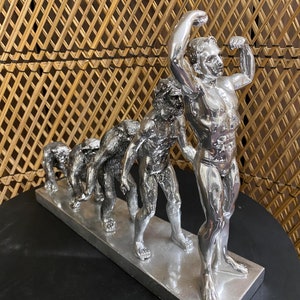 May include: A silver sculpture depicting the evolution of man. The figures progress from ape-like to a muscular human, mounted on a rectangular base. The sculpture is displayed on a black surface with a woven background.