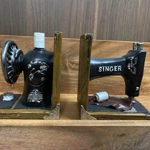 Könnte beinhalten: Ein Paar antike Nähmaschinen-Buchstützen. Die Buchstützen sind aus schwarzem Metall und Holz gefertigt. Die linke Buchstütze zeigt eine Nähmaschine mit einer Garnrolle und einer Nadel. Die rechte Buchstütze zeigt eine Nähmaschine mit einer Garnrolle und einem Stück Leder. Beide Buchstützen tragen den Schriftzug "SINGER".