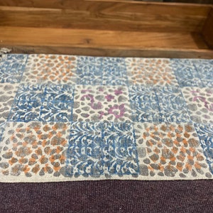 May include: A white cotton rug with a patchwork design featuring blue, orange, and purple floral patterns. The rug has a fringed edge.