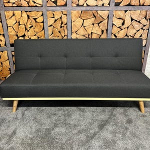 Puede incluir: Un sofá futón de tela gris oscuro con un ribete amarillo claro y patas de madera. El sofá está frente a una pared con un diseño de panel de madera.