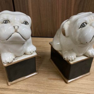 May include: Two ceramic bulldog figurines. The bulldogs are white with black accents and sit atop black rectangular bases with gold trim. The dogs have a slightly grumpy expression. The figurines are decorative and suitable for home decor.