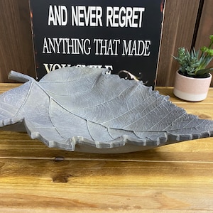 Puede incluir: Un cuenco decorativo de hoja gris hecho de hormigón. El cuenco tiene un diseño de hoja realista con venas y una superficie texturizada.